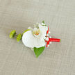 Displayed against a neutral, lightly textured beige fabric that recalls natural linen laid out in a calm Epping townhouse, this boutonnière is centred on a single, flawless white orchid that radiates quiet luxury. Its wide, velvety petals unfurl gracefully, cupping a central column tinged with soft yellow and fine orange speckling, like a tiny flicker of candlelight at the heart of the bloom. Surrounding the orchid, small clusters of white baby's breath create a delicate halo, each miniature flower adding airiness and a whisper of romance that feels perfect for weddings, proms, or black-tie dinners in and around the Epping area. Nestled to the left are two small, bright green buds with a slightly ruffled, mum-like texture, offering a fresh pop of colour and a playful contrast in form. Beneath everything, a smooth, deep green leaf, likely Ruscus, peeks out just enough to frame the orchid and suggest foliage freshly cut that morning by an attentive florist. The stem is tightly wrapped in a striking red satin ribbon, its surface catching the light with a subtle sheen, then finished in a dainty bow that rests on the right side, lending a hint of passion and celebration without overwhelming the design. The composition feels balanced and carefully considered, as if shaped by hands used to dressing lapels for elegant events at local venues near Epping Forest or along the High Street. Though small, the piece suggests a whole story of promise, devotion, and the quiet pride that comes from wearing something beautifully made, close to your heart, on a day that truly matters.