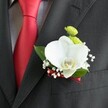 A close-up photograph focuses on a meticulously crafted wedding boutonniere pinned to the lapel of a dark charcoal grey suit jacket, the wool fabric showing a subtle, woven texture and a neatly stitched pocket just visible at the edge of the frame. At the heart of the arrangement sits a single, large white Phalaenopsis orchid, its velvety petals opening in a serene, sculptural curve, with a distinctive yellow-and-white lip drawing the eye like a soft point of light. Nestled around the orchid, delicate clusters of tiny white baby's breath float like a cloud, adding an airy, romantic halo. Just above, two small, round blooms in a vivid lime-green chrysanthemum style introduce a playful burst of colour, their tightly packed petals creating a gentle, cushion-like texture. A few rich green leaves fan out from behind, framing the flowers with a fresh, natural backdrop that suggests the cool greenery of nearby Epping Forest. To the left, a solid red satin tie lies against a pale grey and white small-check dress shirt, the glossy fabric catching the light and echoing the deep red satin ribbon that wraps neatly around the base of the boutonniere. The ribbon is wound with precision, matching the tie in tone and adding a final, sophisticated flourish. Soft, even lighting highlights each petal, stitch, and fold, capturing that poised moment just before the groom steps out for a ceremony in Epping, perhaps at a local church or registry office. The entire composition feels refined yet personal, the kind of thoughtful detail a local florist might create to elevate a classic wedding suit with quiet confidence and enduring style.