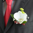 Pinned neatly to a dark lapel, this orchid boutonnière appears like a small, curated garden captured against formal attire, as if the wearer has carried a fragment of Epping Forest to a town hall wedding. At its centre, a pristine white orchid opens in a graceful arc, its petals thick and smooth, sculpted like fine china. A hint of yellow at the throat adds warmth and depth, inviting a closer look. Behind it, two lime-green button chrysanthemums bring a youthful vibrancy, their domed, textured blooms echoing the freshness of early spring along the paths near Bell Common. Clusters of baby's breath scatter around the composition, each tiny white flower creating a constellation of softness that breaks up the darker tones of the suit. Deep green leaves cradle the flowers, adding structure and contrast while subtly guiding the eye downward to the base. There, a neatly wrapped ribbon in a deep, romantic red ties everything together, mirroring the rich red of the tie that sits just below. The interplay of colours-crisp white, vivid green, and luxurious crimson-creates a balanced, sophisticated palette ideal for grooms, best men, or distinguished guests at an Epping celebration. You can almost sense the gentle coolness of the orchid against the fabric, and the quiet rustle of foliage as the wearer moves. The design feels thoughtfully measured, crafted so that no single element overwhelms the others, yet each detail contributes to an overall impression of calm confidence and celebration. It's a modest piece of floral art that turns a simple suit into a carefully composed expression of style and sentiment.
