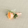 A softly lit photograph showcases a carefully crafted peach rose boutonniere resting horizontally on a pale beige, linen-textured fabric, the fine weave of the cloth echoing the understated elegance of a wedding morning in Epping. At the heart of the design is a single, partially open rose in warm peach tones, its petals shifting gently from a creamy outer edge to a deeper apricot and soft orange at the centre, as if just picked fresh from a sheltered garden near Epping Forest. Around the rose, airy clusters of white baby's breath form a delicate halo of tiny blossoms, their cloud-like texture adding a sense of lightness and romance. Slim, grass-like green sprigs and a couple of broader, darker green leaves curve neatly beneath the bloom, introducing structure and contrast without overwhelming the focal flower. The stems are bound with a smooth, peach satin ribbon, wrapped with the precision of an experienced florist and finished in a small, tidy bow that lies close to the base, ready to be pinned to a lapel. Soft, even lighting highlights the gentle sheen of the ribbon, the velvety surface of the rose petals, and the subtle grain of the woven background, creating a calm, intimate mood. This floral accessory feels ideal for a groom or prom guest heading to a celebration at a local Epping venue, capturing the quiet joy of special occasions and the timeless charm of a classic rose buttonhole tailored for local ceremonies and family gatherings.