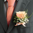 Pinned to the left lapel of a deep charcoal suit, a single peach-tinged rose takes centre stage in a photograph that reads like a small love note. The bloom's petals are layered and nearly translucent in places, softly unfurling to reveal a centre of warm, delicate colour that sits in subtle contrast to the suit's cool tones. Surrounding the rose are sprinkled clusters of white baby's breath-gypsophila-whose tiny, cloud-like blossoms float above narrow sprigs of pale green foliage; these elements lend a delicate, handmade quality and create a gentle frame that lightens the formal silhouette. The boutonnière is neatly finished with a pale salmon satin ribbon, tied low and tidy so its satin glint mirrors the pale peach tie beneath, which rests over a light shirt patterned with a faint dark grid, adding quiet structure. The overall impression is one of considered simplicity and old-fashioned romance-an image that evokes vows exchanged near the tranquil walks of Epping Forest or photographs taken outside the local church on Station Road. You can sense the florist's practiced hand in the balanced proportions and the crisp finishing: a small composition that reads as fresh-cut, subtly scented, and perfectly scaled for menswear. For couples seeking a softened modern classic, this boutonnière suggests calm elegance and thoughtful coordination, a piece made to be noticed in close conversation, in portrait shots on the High Street, or beside family photographs taken after a ceremony in our town.