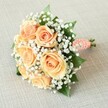 A hand-tied bridal bouquet of soft peach roses and white gypsophila rests on a pale, oatmeal-toned fabric, its rounded shape cradled in gentle, even light that feels like a calm morning in Epping. Around seven or eight roses, fully open or just on the verge of bloom, cluster at the heart of the design, their petals spiralling from tight, warm centres to looser outer curls in shades of apricot, pale orange, and mellow peach. Each bloom looks velvety to the touch, with subtle shading along the petal edges that hints at freshness and careful conditioning. Threaded between the roses are countless tiny stars of white baby's breath, creating a cloud-like halo that softens every line and adds a bright, airy contrast to the warmer tones. Small, glossy green rose leaves peek from within the flowers, adding depth and a natural, garden-grown character reminiscent of roses cut from a well-tended Epping High Street back garden. The stems are neatly gathered and wrapped in a smooth coral-peach satin ribbon, its surface catching the light with a delicate sheen. Tiny white faux pearls, arranged in a gentle spiral along the handle, introduce a refined bridal detail that feels perfect for weddings in local venues around Epping Forest or a ceremony near St John's. The very ends of the cleanly trimmed stems show beneath the wrapping, grounding the elegance with a touch of simplicity. The textured linen- or burlap-like background remains soft and neutral, allowing the flowers to glow as the quiet focal point, ideal for a romantic wedding, anniversary celebration, or intimate registry office ceremony in Epping.