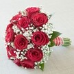 A striking bridal bouquet, expertly crafted by local Epping florists, is presented against a delicately textured pale background, allowing its rich details to command attention. The tightly gathered dome shape showcases an array of fully opened crimson red roses, their velvety petals brimming with lushness and depth. Interspersed among the roses, petite sprays of airy white baby's breath float gracefully, their softness infusing the arrangement with a sense of movement and light. Peeking between the blooms, deep green leaves provide a cool, natural accent, echoing the nearby Epping Forest's tranquility. The handle, meticulously wrapped in wide red satin ribbon and finished with crisp white horizontal stripes and a spiral of pearly beads, feels sumptuous and polished, hinting at the careful attention given in every detail. Fresh green stems emerge neatly at the base, a testament to the bouquet's freshness and quality. Bathed in soft daylight, the bouquet's vivid reds and pure whites seem to glow, the textures and intricate patterns standing out in sharp relief. Perfect for Epping weddings, anniversaries, or special moments of declaration, this bouquet conjures the timeless language of love and celebration-delivering a sense of joy and meaning to any local event.