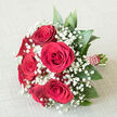 A traditional hand-tied bouquet of sumptuous red roses and airy white baby's breath lies on an off-white, textured cloth, the simple background allowing every detail of the flowers to stand out clearly, like a still-life moment captured in an Epping townhouse. In the centre, a rounded cluster of six to seven deep crimson roses commands attention, each flower fully open yet still perfectly formed, with velvety petals that curl softly at the edges, suggesting both luxury and tenderness. Nestled between and around these roses, clouds of small white gypsophila blossoms scatter like tiny stars, their fine stems weaving through the design to create a light, misty veil that contrasts beautifully with the bold red. Framing the composition, smooth, dark green foliage fans gently outward, adding volume, structure, and a sense of natural ease, reminiscent of the greenery seen along the paths leading into Epping Forest. The stems are firmly bound into a rounded handle, dressed in rich red ribbon that has been carefully wrapped and decorated with a line of tiny white dots or bead-like accents, spiralling downwards to give the impression of delicate jewellery. This elegant handle suggests the bouquet is ready to be carried-perhaps by a bride stepping out of a car near St John's Church, or by someone heading to a romantic dinner at home in Epping. The entire arrangement feels poised between tradition and intimacy, crafted with the quiet confidence of an experienced local florist who understands how red roses and baby's breath can speak of enduring love, commitment, and meaningful celebration.