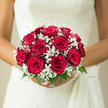 In this image, a bride stands poised, her white dress forming a soft, luminous backdrop for a perfectly rounded bouquet of red roses and baby's breath, as if she were about to step from Epping High Street into a tranquil ceremony space. The roses are the undeniable stars: each one a deep, velvety red, petals layered in tight, confident spirals that create a sense of depth you can almost reach into. Tiny white blossoms of gypsophila weave between and around the roses, creating a ring of airy lightness like a fine lace collar. This baby's breath appears weightless, softening the strong crimson tones and adding a gentle, cloud-like texture that makes the bouquet appear to float above the bride's hands. Beneath the blooms, subtle hints of glossy green foliage peek out, anchoring the design with a touch of nature that evokes the greenery of Epping Forest and nearby country lanes. The bouquet's symmetry and compact, domed form speak of thoughtful craftsmanship and classic bridal style, a choice that feels as appropriate for a ceremony at St John's Church as for an intimate gathering at a local Epping venue. Imagining the scene, you can sense the cool satin of the dress, the faint rustle of tulle, and the soothing fragrance of fresh roses rising with every breath-a blend of sweetness and greenery that calms nervous excitement. This bouquet becomes more than an accessory; it is the bride's quiet companion, held close as she walks down the aisle, smiles at loved ones, and steps into a new chapter of her life.