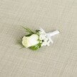A classic white rose boutonniere rests diagonally across a softly textured, linen-like beige fabric, its simple elegance evoking a wedding morning in Epping. At the heart of the design sits a single creamy white rosebud, the petals still tightly furled yet velvety and full, hinting at the moment it will just begin to open. The bloom is clean and luminous, with a faint blush of ivory in the shadows and a tiny glimpse of green sepals cupping the base. Around it, clusters of airy white baby's breath form a gentle halo, each tiny flower like a floating snowflake, adding a cloud-soft texture that makes the rose appear even more pristine. Slender, deep green leaves and a few feathery sprigs of foliage peek from behind the petals, framing the boutonniere with natural depth and a fresh garden feel, as though the stems were just gathered from a quiet corner of an Epping garden near the High Street. The stems are neatly bound in a narrow band of pure white satin ribbon, carefully wrapped to create a smooth, polished handle that sits comfortably in the hand. Just below the bloom, the ribbon is tied into a small, perfectly formed bow, its tails lying flat and tidy, suggesting the steady hands of an experienced florist preparing it for a groom's lapel. Soft, diffused light washes over the arrangement, highlighting the satin sheen of the ribbon, the gentle curve of each rose petal, and the subtle weave of the fabric background. The overall impression is of calm sophistication and quiet anticipation, an understated boutonniere suited to a traditional ceremony at a local church or registry office, capturing themes of purity, new beginnings, and refined wedding style in Epping.