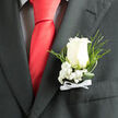 Pinned neatly to the breast of a deep charcoal suit, this classic white rose boutonniere is photographed in intimate detail, showing every curve and fold of its velvety petals. The rose itself is a soft ivory-white, the outer petals just beginning to relax open, revealing a calm spiral at the heart that feels both timeless and serene. Around the base of the bloom, clusters of baby's breath form a halo of tiny, snow-like flowers, their minute blossoms adding texture and a sense of lightness. Behind and around them, slim arcs of fresh green foliage and feathery fern fronds extend outward, giving the arrangement a natural, slightly flowing silhouette that contrasts with the clean lines of the suit. The stems are bound together with a narrow white ribbon, tied into a compact bow that sits just above the pocket, a subtle nod to tradition and care. The charcoal lapel is crisply tailored, the fine weave of the fabric catching a gentle indoor light, echoing the polished feel of a wedding at a country hotel near Epping or a ceremony in a church tucked off the High Road. The overall mood is one of composed joy - the kind of understated elegance chosen by a groom or best man who values simple, meaningful details. In this image, you can almost imagine the faint, fresh scent of the rose mingling with the cool evening air as guests gather from nearby areas like Buckhurst Hill and Chigwell, pausing to notice this small but significant floral touch.