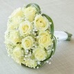 Viewed closely atop a light beige fabric, this elegant bouquet captures the artistry of Epping florists and the spirit of classic celebrations. At its heart, a tightly packed circle of a dozen creamy white and pale yellow roses showcases petals that glow softly beneath diffused daylight-each rose a testament to freshness and subtle beauty. Between the roses, generous clouds of tiny, snow-white gypsophila add a feathery, ethereal quality, as if morning mist had settled gently among the blooms. Loops of lustrous, dark green foliage curl gracefully around the bouquet, their deep tones contrasting with the delicate florals and framing them in rich color. The stems are meticulously gathered and wrapped in a white satin ribbon, forming a smooth, dignified handle ready for a bride or to be presented at a milestone celebration. The backdrop-a softly textured linen-hints at special occasions across Epping, from wedding mornings in Bell Common to romantic gestures between friends near Copped Hall. Lush, fresh, and romantic, this arrangement conveys enduring love and new beginnings, a perfect illustration of Epping's tradition of elegant floral design.