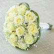 There is an almost dreamlike hush to this bridal bouquet, captured from a slightly raised viewpoint so the dome of flowers reads like a soft, luminous halo. Nestled at its centre are about fifteen to twenty creamy-yellow roses, their petals spiralling to flawless hearts that glow in soft, even light. Between them, fine sprays of white Gypsophila breathe air and delicacy into the composition, creating pockets of airy contrast that let each rose speak. Curving around the outer edge, a few long, vibrant green blades-bear grass or lily grass-arch with gentle, calligraphic lines, embracing the blooms and adding a contemporary silhouette to a classic form. The natural stems are visible beneath a pristine white satin wrap, wound with care into a cool, comfortable handle. The bouquet sits on a pale, slightly textured linen that tones the cream and green into an elegantly muted palette, the kind of surface we use in the Flowers Epping studio when preparing orders for nearby ceremonies. The lighting is soft and diffused, emphasising slight variations in the roses' pale yellow and the satiny sheen on the ribbon. Close your eyes and you'll imagine the scent: a soft, understated sweetness with green hints, like early morning strolls under Epping Forest trees. This arrangement feels made for intimate weddings, small registry celebrations, or anniversary surprises delivered along the High Street or to homes around Theydon Bois-an emblem of refined restraint, quietly joyful and immaculately crafted by a local hand.