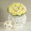 Creamy yellow roses gather into a perfectly rounded dome, their layered petals unfurling with a softness that feels both cheerful and composed. Interspersed throughout are clouds of white gypsophila; the minute blossoms float like notes of breath, softening the bouquet and lending a delicate halo against the roses' warm tones. The flowers are seated in a contemporary white-washed wicker orb, its open, sculptural weave creating negative space that makes the florals appear to hover-an effect I often favour when styling arrangements for intimate gatherings in Epping. Curving green blades weave around the arrangement's edge, their gentle loops adding a rhythm that contrasts the roses' solidity. Nearby, small woven spheres introduce a playful, minimalist accent, their matte texture mirroring the basket and offering a tactile counterpoint to the petals' silky sheen. In bright, natural light the lemon-cream colour of the roses becomes almost luminous, and there is a subtle fragrance: a clean, fresh-cut meadow note with a whisper of earth that grounds the sweetness. This composition reads as hopeful and serene, equally suited to a congratulatory gift collected after a stroll through Epping Forest or as a restrained centrepiece at a family celebration on the High Street. Prepared with considered balance and technical care, the bouquet reflects neighbourhood sensibilities-fresh, restrained and timeless-and, for customers close by, Flowers Epping can tailor similar designs for local hand-delivery with that same attentive finish.