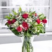 A lush, romantic bouquet of red roses, ruffled red carnations, white alstroemeria, and airy baby's breath stands proudly in a clear glass vase on a bright white tabletop, bathed in soft natural light from a nearby window in Epping. The deep crimson roses are the stars, some fully open with velvety petals spiralling outward, others still in elegant bud form, promising days of unfolding colour and scent. Nestled amongst them, plump carnations in the same rich red add volume and a plush, almost cushiony texture, giving the arrangement a traditional yet timeless charm. Clusters of white alstroemeria, some still in tight green-tipped buds, weave between the reds, their partially open blooms showing slender, trumpet-shaped petals that feel fresh and hopeful, like early morning in Stonards Hill Park. Delicate sprigs of gypsophila, or baby's breath, drift throughout like a soft, white mist, creating a cloudlike halo that lightens the overall composition and gently diffuses the bold tones. Around the base, large, glossy green leaves cradle the flowers, forming a structured collar that both grounds and elevates the bouquet, while finer, lighter foliage threads through the centre to add movement and depth. Through the clear glass, neatly trimmed stems are visible, immersed in clear water that glints faintly with the light, underscoring the freshness of a bouquet just prepared by an attentive florist. Behind the arrangement, the outside world is reduced to a soft, grey blur through the window, making the vivid reds, whites, and greens appear even more intense. The scene feels like a quiet moment of celebration, perfect for an anniversary dinner on a quiet Epping High Street evening, a heartfelt thank-you, or simply to brighten someone's home with an elegant note of love and appreciation.