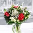 A contemporary bouquet of red and white flowers stands in a clear glass vase, arranged with the care of an experienced Epping florist and lit by soft daylight streaming through a nearby window. At the heart of the design, a single coral-red rose and a plush crimson carnation draw the eye, their velvety petals layered and full, like rich fabric gathered into delicate folds. Around them, clusters of dainty white chrysanthemums and tiny daisy-like blooms form a gentle halo, their crisp petals fanning out like little stars against the deeper colours. Creamy white lilies, some fully open and others still in smooth, pointed buds, rise gracefully from the centre, adding height and a sense of anticipation, as if the bouquet is still quietly unfolding. The entire arrangement is cradled by broad, glossy emerald-green leaves, skillfully folded and curved into sweeping arcs that hug the base and sweep upwards, giving the bouquet a sculptural, modern silhouette. The foliage has a polished sheen that catches the light, creating subtle reflections on the water in the vase and on the table beneath. The mood is uplifting yet calm, like a cool, clear morning walk through Epping Forest or along the High Street before the shops have fully opened. You can almost imagine the faint, fresh fragrance of lilies mingling with the leafy green scent of the stems, a whisper of spring even on a grey Essex day. This is the sort of arrangement that would feel perfectly at home brightening a kitchen in Theydon Bois, celebrating a birthday in Loughton, or simply adding a thoughtful touch of beauty to an everyday moment at home in Epping.