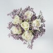 An overhead, high-angle photograph shows a compact, circular bouquet laid on a pale off-white surface, its simplicity allowing every detail of the flowers to stand out. At the very heart of the design are six creamy white roses, each bloom tightly packed with velvety petals that curl in perfect spirals, like small ivory sculptures. The roses are unblemished and serene, with the faintest touches of pale green at the base of a few outer petals and sepals, hinting at their freshness as if they were cut only moments ago from a garden near the edge of Epping Forest. Surrounding these central blooms is a soft, cloud-like ring of lilac and muted lavender filler flowers, likely statice or limonium, whose countless tiny petals create an airy, feathery halo. Their delicate purple tones are gentle rather than loud, bringing a soothing contrast that makes the white roses glow even more brightly. The bouquet is carefully shaped into a full, rounded dome, suggesting the steady hands and practiced eye of a local florist used to preparing bridal posies and sympathy tributes for families across Epping High Street and the nearby lanes. Soft, even lighting washes over the flowers from above, revealing the subtle texture of each petal and the fine, papery quality of the statice, without any harsh shadow to disturb the calm. The mood is quiet and contemplative, ideal for a wedding, a thoughtful anniversary surprise, or a comforting gesture sent to a home near Bell Common, conveying purity, remembrance, and gentle affection in a way that feels timeless and deeply personal.