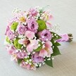 Resting softly against a subtle, cream-toned background, this vibrant hand-tied bouquet exudes joy and romance-an artisan design inspired by the changing seasons and community spirit of Epping. At the heart of the arrangement, plush roses shimmer in tones of lilac and pink, their layered petals unfurling elegantly like a blush at first light over Epping Green. These classic blooms are flanked by cheerful pompom chrysanthemums in brilliant lime green, lending the piece a bold, refreshing contrast as lively as the bustling High Street on market day. Blush-pink and pale lily-like flowers-delicate Alstroemeria-add graceful curves and streaks of maroon and yellow, weaving movement and visual poetry through the cluster. Threaded throughout, clouds of gossamer white baby's breath soften and unite the vibrant palette, imbuing the bouquet with a gentle, ethereal charm. Lush green leaves peek from between blossoms, creating balance and depth. The stems are carefully aligned and wrapped in a gleaming pink ribbon, secured with pearl accents, ready to be held by a bride, gifted for love, or placed as a centrepiece at a celebration on Epping's leafy outskirts. Each element speaks to the freshness, artistry, and meticulous attention of a skilled local florist, making this bouquet a fitting tribute to cherished moments in Epping.