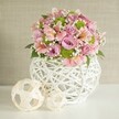 Brimming with natural grace, this floral centerpiece brings a touch of handcrafted luxury to any setting, echoing the elegant simplicity beloved by Epping's residents. Resting on a spotless white surface, the bouquet is composed of lush, open roses in pastel pink and lavender, flanked by clusters of vivid lime green button chrysanthemums for added cheer. Delicate pink alstroemeria lilies, easily identified by their unique striped markings, contribute both a romantic flair and a hint of exotic interest. Interlaced throughout, wispy white sprigs of baby's breath lend an ethereal softness that makes the arrangement feel as welcoming as a friendly greeting on Epping High Street. The composition is cradled in a contemporary white vase, its rounded form fashioned from interwoven strips that create an open, basket-like appearance-blending rustic craftsmanship with fresh, airy design. Beside the vase, two decorative spheres made of pale, woven material reinforce the craft motif, their subtle presence balancing the scene. Gentle daylight washes over the ensemble, enhancing every color and highlight. Designed with an experienced local florist's eye, this image embodies the spirit of celebration and personal connection-perfect for weddings at Theydon Bois or as a chic centerpiece gracing a family table near Epping Forest, where flowers are cherished for marking life's most beautiful moments.