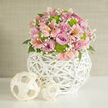 A stylish, rounded bouquet sits on a pristine white tabletop, bathed in gentle daylight that seems to stream in from an unseen window, like a bright morning in an Epping townhouse. The arrangement is densely packed yet carefully balanced, with pale lavender and deeper pink roses forming the main body, their petals unfurling in soft, layered spirals. Nestled among them are graceful stems of light pink alstroemeria, each blossom showing fine speckles and streaks on its inner petals, as if hand-painted. Small, domed clusters of vivid lime-green button chrysanthemums pop out between the pastels, bringing a crisp, modern twist and adding a pleasingly firm texture. Delicate sprays of white baby's breath weave through the bouquet, their tiny blossoms creating a halo effect that diffuses the light and gives the flowers a dreamy softness. Hints of dark green foliage appear around the base, just enough to frame the blooms without overwhelming them. The bouquet is held in a globe-shaped vase crafted from white woven rattan or wicker, its open lattice pattern forming an airy, sculptural cradle that feels both contemporary and welcoming. Nearby, two smaller decorative spheres in a lighter, natural weave echo the vase's design, adding a relaxed, homely detail that would suit a wedding display in a local Epping venue or a stylish dining table off the High Street. The background is a simple, neutral-toned wall, ensuring the eye stays on the arrangement, which exudes a mood of gentle celebration, fresh beginnings, and thoughtful design.