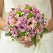 In this image, a romantic hand-tied bouquet is presented as if ready to be carried from an Epping doorway straight to a wedding aisle. At its centre, dusky mauve and lilac roses form a calm, elegant core, their petals spiralling inward in perfect rosettes that look soft and cool to the touch. Surrounding them, pink alstroemeria stretch outward in graceful arcs, each bloom dotted with fine speckles and streaks of deeper colour, like brushstrokes on a watercolour painting. Between these larger flowers, bright green button chrysanthemums stand out in rounded clusters, their chartreuse hue adding a lively spark that keeps the palette from feeling too demure. Fine sprays of gypsophila drift through the bouquet, their tiny white blossoms filling the gaps with a gentle cloud-like haze, as delicate as whispered vows exchanged in a registry office near Epping High Street or a private garden celebration. Glossy greenery anchors the arrangement, providing contrast and depth; some leaves curve protectively around the blooms, while others peek through, catching the light and reinforcing the sense of freshness. The bouquet is photographed in soft, even daylight, allowing the lilac, pink, and green tones to glow naturally without harsh shadows, as if positioned near a bright window in a local florist's studio. The stems are tidily gathered, suggesting a comfortable, balanced grip that would feel secure during a long day of photos, toasts, and dancing. The overall impression is one of quiet, joyful elegance-a carefully composed wedding bouquet that celebrates partnership, tenderness, and hope, ready for same-day delivery to couples planning their special moments across Epping and nearby villages.