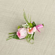 This image shows a carefully composed wedding boutonniere centred on a cool, soft pink rose, its petals layered like satin and curling gently outward, each edge catching a line of light. Around it, pastel alstroemeria blooms provide a delicate chorus, their petals tinged with blush pink and streaked subtly with deeper lines that draw the eye towards their creamy yellow centres. Threaded between them are tiny white gypsophila blossoms, each one a minuscule puff of texture, creating a halo that softens the overall shape. Slender sprays of greenery, fresh and slightly glossy, arc gracefully around the flowers, their thin stems and narrow leaves adding movement and contrast. The entire cluster is bound at the base with a smooth, pink satin ribbon, wrapped tightly and finished with expert precision, the sheen of the fabric mirroring the rose's gentle glow. Beneath the arrangement, a piece of lightly textured fabric acts as the backdrop-perhaps a tablecloth in an Epping wedding venue or the work surface in a local studio-its neutral tones ensuring the floral colours stand out with quiet elegance. The lighting feels natural, like daylight filtering in from a nearby window along Epping High Street, giving the rose a softly luminous quality without harsh shadows. You can almost imagine the crisp feel of the greenery, the velvet softness of the rose, and the faint, fresh fragrance that would rise when this boutonniere is pinned to a lapel. It's a piece created for significant moments-a groom at a countryside ceremony, a prom guest stepping out in Epping-expressing tenderness, care, and understated sophistication.