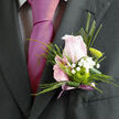 The image captures a refined boutonnière, carefully composed and ready to be pinned to a lapel for a wedding or formal event in Epping. At its centre is a tightly formed blush-pink rose, its petals smooth and velvety, opening just enough to hint at the fullness still to come. Surrounding the rose are cool, textural greens: a folded leaf that frames the bloom with clean lines, small sprigs of foliage, and a single elongated blade that extends upward, introducing a sleek, modern silhouette. Tiny white gypsophila, arranged in dainty clusters, softens the structure, their minuscule blossoms resembling floating flakes of snow against the darker greenery. A small green chrysanthemum button flower is tucked near the base of the rose, its concentric petals and vivid colour lending a playful yet sophisticated contrast. The stems are bound together neatly, then finished with a slender ribbon in muted lavender tones, the bow lying flat and discreet so as not to overpower the flowers. The lighting is gentle, highlighting the natural textures of the petals and leaves and casting delicate shadows that enhance the depth of the piece. It is easy to imagine this boutonnière being delivered fresh from a local florist's workshop just off Epping High Street, carried carefully to a groom waiting at the registry office or a charming country venue on the edge of Epping Forest. The overall mood is one of subtle celebration - an understated, handcrafted detail that quietly speaks of care, artistry, and heartfelt preparation.