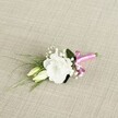 A delicately crafted wedding boutonnière lies diagonally across a softly textured, light beige fabric that looks like finely woven linen, its natural oatmeal tone reminiscent of the calm, neutral interiors of homes around Epping High Street. At the heart of the design sits a fully open white lisianthus bloom, its ruffled petals layered in gentle curves, creating a soft, cloud-like centrepiece that suggests purity and quiet celebration. Just to the left, two slender, cream-tinted lisianthus buds rest closely together, their pale green tips still tightly closed, capturing the tender promise of a moment not yet fully unfolded, like a couple on the morning of their wedding at a nearby Epping church. Surrounding these focal blooms, tiny sprays of white gypsophila weave in and out, each miniature flower catching the light like delicate drops of dew, adding a romantic, airy fullness to the small arrangement. Wisps of fine, grass-like greenery arc gracefully towards the lower left corner, their linear shape giving the piece a sense of movement and natural flow, as if caught in a gentle breeze blowing across Epping Forest. Deeper green leaves, broader and more structured, nestle behind the white petals, providing a lush backdrop that frames the central flower and enhances its bright clarity. At the base, the slender stems are neatly gathered and wrapped with a vibrant fuchsia satin ribbon, tied into a small, jaunty bow whose silky sheen offers a cheerful pop of colour against the muted background. The overall look is elegant yet modern, perfect for pinning to a lapel for a wedding, prom, or formal celebration anywhere in and around Epping, capturing the refined charm of a thoughtfully chosen floral detail.
