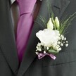 A sharply focused close-up of a groom's charcoal grey suit jacket, captured just at the lapel so the textures and florals feel almost touchable, as if you were standing with him outside the registry office in Epping High Street. The fine wool suit has a subtle woven pattern, its right lapel crisply pressed and catching soft, even light. Between the lapels sits a smooth, perfectly knotted silk tie in vivid magenta or deep fuchsia, glowing against a pale grey and white patterned dress shirt. Pinned to the left lapel is a carefully crafted buttonhole: a single fully opened white lisianthus, or rose-like bloom, its many ruffled petals layered into a soft, cloud-like centre. Just above, two slender ivory buds lean gently forward, still closed and full of promise, framed by airy strands of feathery green foliage. Baby's breath, tiny and snow-like, clusters around the main flower, adding a light, romantic halo that softens the line of the jacket. At the base, a broad, glossy green leaf curves protectively around the stems, neatly wrapped and finished with a narrow mauve ribbon that echoes the tie's rich tone. The ribbon is tied with meticulous precision, suggesting a florist's practiced hands and the quiet anticipation of a wedding morning. The composition feels calm yet celebratory, ideal for describing formal men's wedding attire, groom or groomsmen styling, or same-day wedding flower delivery for ceremonies around Epping Forest, Theydon Bois, or nearby village churches, while remaining fully focused on the visible details of the suit and boutonnière.