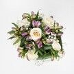 A full, hand-tied bouquet is photographed directly from above against a bright, seamless white background, so every bloom appears crisp and luminous. At the heart of the arrangement, creamy white and soft ivory roses cluster together, some still cupped in tight buds with a faint buttery-yellow glow at their centres, others fully opened to reveal spiralling layers of velvety petals. Between the roses, stems of alstroemeria add gentle drama: slender buds and half-open flowers show off petals washed in mauve, lavender, and purplish-pink, with fine green streaks that echo the freshness of early spring in Epping's High Street gardens. Sprays of tiny white gypsophila thread through the bouquet like drifting clouds, softening the edges and creating a misty, romantic halo around the larger blooms. Beneath and around everything, rich deep-green foliage and broad, glossy leaves cradle the flowers, giving the design a compact yet generous shape, like a cupped armful of blossoms just gathered for delivery to a door off Station Road. The light is clean and even, with no harsh shadows, highlighting the textures of each petal and leaf and conveying a sense of cool, tranquil elegance. The overall feeling is of quiet celebration and refined affection, an arrangement that could easily be imagined on a wedding table in a country house near Epping Forest, or as a thoughtful gift to brighten a calm corner at home. The bouquet feels meticulously composed yet natural, a study in soft whites and greens accented by gentle lilac tones, captured in a single overhead moment of still, graceful beauty.