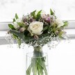 An elegant white and green bouquet sits in a clear cylindrical glass vase, poised in front of a softly glowing white window that blurs gently into the background, like a bright Epping morning mist over the fields near Bell Common. At the heart of the arrangement, three fully open white roses with creamy, velvety petals and subtle ivory centres form a serene focal point, their layered petals unfurling in perfect spirals that convey purity and quiet confidence. Around them, lush green foliage in varying shapes and textures is carefully structured: broad, folded leaves curve protectively around the roses, creating an emerald cradle, while slimmer stems add height and a gentle sense of movement, as if stirred by a light breeze drifting up from Epping Forest. Threaded delicately through the design, tiny white gypsophila blossoms - baby's breath - scatter like soft clouds or floating confetti, lending an airy, romantic haze that lightens the whole composition. Nestled between the greenery, alstroemeria buds and blooms introduce hints of muted mauve and pale blush-pink, their slender petals and faint streaks of colour suggesting new chapters just beginning to open. The stems are visible through the transparent vase, clean and straight, gathered neatly and secured with a fine ribbon just above the waterline, the clear water catching the light. The overall shape is compact and rounded, a classic, balanced design that feels thoughtful and composed, ideal for a peaceful wedding at a local Epping venue, a simple thank-you delivered to a High Street home, or a gesture of gentle comfort. The bright, natural light bathing the bouquet enhances every texture and tone, wrapping the scene in a tranquil, almost meditative glow.