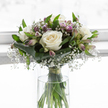 A tranquil white and soft-lilac bouquet stands poised in a simple glass vase, the flowers bathed in calm, diffused light that suggests a peaceful room somewhere off Epping High Street or overlooking the edge of Epping Forest. At its core, fully opened white roses sit in gentle prominence, their petals thick, velvety, and perfectly layered, each bloom forming a rounded cup that conveys tenderness and care. Around them, tiny flowers of white baby's breath cluster like fine mist, filling the gaps with a dreamy, cloud-like softness that lightens the entire composition. Among these, slender alstroemeria stems appear, some still held in bud, others just unfolding into narrow petals shaded in lavender, soft purple, and delicate stripes, adding a quiet hint of colour that speaks of remembrance and hope. Encircling the flowers, broad, deep green leaves are wrapped and folded with precision to create a smooth collar, while narrower leaves weave upward between the stems, suggesting life and continuity. The clear cylindrical vase reveals the straight, fresh green stems gathered together, their base submerged in clear water and tied just above the surface with an unobtrusive green band to hold the bouquet's classic rounded shape. The minimal, light background keeps all attention on the blooms themselves, amplifying the sense of purity and stillness. This arrangement feels especially suited to sympathy occasions in the Epping area-resting beside a candle during a memorial, placed in a quiet hallway, or delivered to a grieving family as a gentle, living gesture of comfort, respect, and enduring affection.