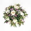 An overhead, top-down view shows a perfectly rounded hand-tied bouquet laid against a crisp white background, the clean space making every petal stand out in vivid detail. At the heart of the design, creamy white roses in shades from soft ivory to pale champagne spiral open, some fully unfurled and plush, others still cupped and expectant, their velvety petals suggesting a gentle, powdery fragrance. Threaded between these classic blooms are clusters of Alstroemeria buds in deep lavender and rich purple, their tips brushed with fresh green as if just picked from a garden near Epping Forest. The buds are mostly closed or only slightly parted, adding a sense of quiet anticipation and texture, their narrow petals streaked with fine, painterly markings. Tiny clouds of white gypsophila drift through the arrangement, each miniature blossom no bigger than a pinhead, creating a soft, misty veil that lightens the richer tones. Around the edge, glossy, dark green leaves frame the flowers like a natural wreath, the foliage layered and slightly overlapping, giving depth and a sense of movement. Soft, even daylight reveals the subtle curves and shadows of each bloom without harsh contrast, evoking the calm mood of a bright but peaceful morning on Epping High Street. The bouquet feels balanced yet alive, poised between formality and tenderness, ideal for sending comfort, marking an anniversary, or simply bringing a sense of hope and renewal into a home in CM16 and the surrounding lanes.
