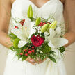 A close view of a hand-tied bridal bouquet rests against a softly patterned lace wedding dress, the intricate fabric framing the flowers like a delicate backdrop. Dominating the composition are elegant white lilies, their broad, satin-like petals fully open, with a gentle cream glow at the centre that gives them depth and warmth. Nestled among them is a single, lush red rose, its velvety texture and rich hue speaking of devotion and heartfelt emotion. Surrounding these focal blooms are airy sprigs of baby's breath, each tiny white flower adding lightness, as though a gentle mist has settled across the arrangement. Red berries dot the bouquet with cheerful bursts of colour, while mossy green cushions and small tufts of dianthus bring in a natural, almost woodland texture that recalls the greenery around Epping Forest or the paths near Bell Common. Fine blades of grass and gently arching foliage extend outward, lending a sense of movement, as if the bouquet is lifting slightly from the bride's hands. The light appears soft and natural, perhaps from a window in a quiet Epping venue, making the whites appear luminous and the reds deep and romantic. Every stem seems carefully placed, with an artisan's eye for proportion and harmony. The bouquet feels both luxurious and grounded in nature, like something a local Epping florist might design for a bride who wants her wedding flowers to mirror the calm beauty and fresh air of the surrounding countryside, while still honouring the timeless tradition of red roses and white lilies.