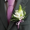 A close-up, high-angle photograph shows the upper torso of a man dressed for a formal occasion in Epping, his dark charcoal grey suit jacket filling most of the frame. The camera lingers on the left lapel, where a meticulously crafted wedding buttonhole brings a soft burst of colour against the structured fabric. The suit is sharply tailored, with a clean shoulder line and a neatly pressed, pale dress shirt beneath, patterned with a faint grid that catches the light. Down the centre runs a smooth, solid lavender tie, its satin sheen echoing the romantic tones of the floral work. Pinned precisely to the lapel is the boutonniere itself: at its crown stands a slender spike of pale green buds, likely hyacinth or Muscari, their tight formation creating a sense of fresh anticipation, like spring along the paths of Epping Forest. Nestled at mid-height are white alstroemeria blooms, their petals creamy and open, each one brushed with delicate streaks of maroon or deep plum and revealing warm golden stamens at the heart. Around them, tiny clouds of white gypsophila hover, the baby's breath softening any hard edges and lending a dreamy, airy quality. Fine, grass-like blades arc gently outwards, while broader, deeper green leaves frame the composition and intensify the contrast against the dark wool suit. At the base, a small lavender satin bow, perfectly matched to the tie, gathers the stems into a tidy cluster, suggesting careful local craftsmanship by an Epping florist. The overall impression is one of poised elegance and quiet emotion, perfectly suited to a groom or groomsman ready to step into a church or country venue nearby.