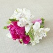 A compact, hand-tied floral arrangement lies horizontally on a softly textured, light beige fabric that looks like natural linen, the neutral background allowing every petal and ribbon to stand out. At the heart of the design is a lush fuchsia peony-style bloom, its many ruffled petals cupped tightly together to create a rich, rounded focal point that seems almost velvety to the touch. Clustered around this vibrant centre are crisp white alstroemeria, their slender, trumpet-shaped petals gently reflexed and brushed with fine maroon streaks and speckles deep in the throat, like delicate ink strokes. Slim, elongated green buds and spiky florets thread through the flowers, adding a fresh, linear texture and a lively contrast of bright green against the pinks and whites. The stems are gathered neatly and wrapped in a glossy ribbon of vivid magenta satin, the smooth sheen echoing the intensity of the central bloom and giving the piece a polished, celebratory finish. Soft, even lighting from above casts gentle shadows into the folds of the petals and along the linen weave, enhancing depth without harshness, as if the piece is resting on a table in an Epping High Street studio just before being pinned on for a summer wedding or prom. The overall impression is of an elegant boutonnière or small corsage, freshly made, with a romantic yet modern colour palette that would suit weddings near Epping Forest, intimate anniversaries at home, or special evenings out in the local village, capturing a moment of joy in a small, unforgettable cluster of flowers.