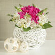 A compact, artfully designed bouquet nestles inside a crisp white wicker basket, its shape perfectly round and pleasingly symmetrical, like a small floral sculpture placed on a bright windowsill in an Epping townhouse. At the centre, a vivid cluster of fuchsia blossoms forms a bold heart of colour, the petals densely layered and velvety, suggesting depth and abundance. Surrounding them, creamy ivory alstroemeria fan outward in a soft halo, each blossom delicately streaked and speckled, their petals curling slightly at the edges as if caught mid-sigh. Between the flowers, slender green stems and glossy leaves introduce a gentle rhythm, guiding the eye around the arrangement. A few buds remain tightly closed, tipped with fresh green, hinting that the display will continue to evolve over the next few days, much like the changing seasons across Epping's forest paths. The basket itself, woven from immaculate white strands, has a modern yet timeless feel, its lattice pattern echoing the light and shadow that fall across the ensemble. On the smooth white surface beside it rest two cream, handwoven spheres, subtle accents that mirror the basket's craftsmanship and add an extra touch of design-forward charm. Soft, filtered light, similar to an overcast spring afternoon near Stonards Hill, bathes the flowers, enhancing their natural colours without harshness and creating a tranquil, uplifting atmosphere. This arrangement feels ideal for marking a fresh start-a small wedding celebration, an engagement dinner at home, or simply a thoughtful delivery to a loved one in Epping who appreciates understated beauty and meticulous floral artistry.