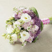 A refined hand-tied bouquet lies horizontally on a pale beige, lightly textured fabric backdrop, the softness of the cloth echoing the gentle mood of the flowers. Dominating the arrangement are lush white lisianthus, their multi-petalled blooms opening like silk rosettes, some revealing subtle yellow-green throats, others still enclosed as promising buds. Between them, pastel pink and lilac-mauve stock flowers form generous, feathery clusters, each floret ruffled and delicate, contributing both volume and a dreamy, romantic haze of colour. Scattered throughout are striking purple filler flowers, reminiscent of ornamental allium, with small star-shaped blooms gathered into spiky globes that punctuate the bouquet with lively lavender-purple energy. Long, narrow strands of glossy green foliage sweep and curl around the arrangement, particularly along the right edge, adding a sense of rhythm and gentle movement, much like the breeze that runs across Epping's open edges of the forest. The stems are cleanly aligned and bound with a vibrant fuchsia satin ribbon, tightly wrapped for a comfortable grip, and finished with a fine string of tiny white pearls that glimmer subtly, giving the handle an almost bridal jewellery feel. This thoughtful blend of soft whites, blush tones, and vivid purples feels ideal for a wedding bouquet or special occasion posy, ready to be carried from a home near Epping High Street to a ceremony in a nearby church. Crafted with the eye of an experienced local florist, it conveys romance, elegance, and the quiet excitement of a meaningful celebration shared close to home.