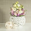 A softly lit floral still life shows a generous, pastel bouquet arranged in a contemporary white sphere vase, set on a crisp white tabletop that could easily belong in a calm Epping living room. The arrangement is abundant with creamy white and pale lemon eustoma, or lisianthus, their many ruffled petals opening like tiny layered gowns. Threaded between them are stocks in gentle shades of blush pink and lilac, their clustered florets rising slightly taller and adding a vertical rhythm, much like the flowering borders you might see along Bell Common in late spring. At the base, a halo of deeper purple filler blooms, perhaps aster or small allium heads, forms a rich, textured collar, framed by a neat ring of deep green foliage that quietly grounds the design. The flowers spill effortlessly from a round, open-work vase created from interlaced white rattan or painted twigs, its airy, basket-like lattice revealing glimpses of the inner container and echoing modern, Scandinavian-inspired decor often chosen in new Epping homes. To the left, two decorative spheres made from pale natural wood or rattan sit companionably on the tabletop, their woven, open patterns mirroring the texture of the vase. Behind everything, a warm greige wall with a subtle vertical fabric or wallpaper grain adds gentle depth without stealing attention, allowing the soft yellows, whites, pinks, and purples to glow. The whole scene feels serene and welcoming, like a thoughtfully styled centrepiece for a quiet birthday breakfast or a relaxed anniversary dinner at home, and perfectly reflects the kind of airy, natural arrangements often requested for same-day delivery around Epping High Street and the surrounding lanes.