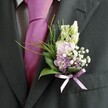 A detailed close-up shows the upper body of a man in a sharply tailored charcoal suit, the kind you might see on a groom arriving at an intimate wedding near Epping High Street. The camera focuses on the left lapel of his jacket, where a carefully crafted buttonhole sits just above a crisp, pale dress shirt woven with a subtle check pattern. A bold magenta-pink silk tie runs down the centre of his chest, its smooth, lustrous surface catching the soft ambient light and echoing the joyful mood of a celebration at one of the barns on the edge of Epping Forest. Pinned neatly to the lapel, the boutonnière is a small work of floral art: slender, unopened buds of creamy white blooms rise at the top, while below them a cluster of soft pink and light lavender flowers-reminiscent of sweet peas or dainty stock-spill gently outward. Tiny clouds of white gypsophila are threaded between the petals, their airy dots lightening the composition, while a few slightly deeper purple florets add satisfying depth. At the base, glossy dark green leaves cradle the flowers, edged with fine, feathery sprigs of bright green foliage that arc outwards like miniature ferns. A lavender satin ribbon is tied in a tidy bow around the stems, its sheen subtly picking up the lilac tones in the flowers and complementing the strong pink of the tie. The overall impression is of a thoughtfully coordinated buttonhole, fresh and elegant, captured in natural daylight and ready to be worn by a groom or honoured guest at a refined Epping wedding. The textures-silk, soft petals, and structured greenery-combine to create a sense of quiet confidence, romance, and careful local craftsmanship.