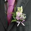 Pinned neatly against a dark, tailored suit, this boutonnière offers a gentle whisper of romance rather than a shout, the kind of refined detail you might see at a wedding reception tucked away near Epping Forest. At its heart, pale lavender blossoms unfurl with soft, velvety petals, their tones hovering between lilac and dusky mauve, conjuring the impression of early spring evenings along Epping High Street when the last light lingers in the sky. Surrounding these central blooms, frothy white filler flowers-dainty gypsophila-scatter like tiny stars, filling the small arrangement with airy volume and a cloudlike texture. A ring of polished, deep green leaves cradles the flowers, their curved shapes framing the pastel centre and lending a grounded, natural feel, as though the stems have only just been gathered from a local garden. Fine sprigs of grass and light, feathery greenery peek out and curve upward, adding a slightly untamed edge that keeps the design from feeling too formal or stiff. At the base, a sheer lavender ribbon is wrapped smoothly around the gathered stems, its translucence allowing hints of green to show through while echoing the colour of the flowers and the coordinating tie beneath. The satin knot lies flat and secure, giving the impression of careful, unhurried preparation before the ceremony begins. This small floral piece feels intimate and personal, an emblem of grace and affection carefully composed by an experienced florist in Epping-perfect for a groom, best man, or cherished guest who wants to wear something that speaks softly of love, craftsmanship, and the promise of a joyful celebration.