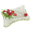 A solemn, handcrafted sympathy pillow formed as a softly rounded rectangle, photographed against a clean white background. The entire surface is densely carpeted with hundreds of small, pristine white chrysanthemum blooms whose tightly layered petals create a pillowy, fleece-like texture that suggests calm and repose. Around the gently raised edges a thin band of pale lime-green ribbon or foliage peeks through, giving a delicate frame to the cushion. On the upper-left corner a concentrated spray of deep red blooms - velvety roses in various stages from tight bud to full bloom, and rich burgundy carnations with their ruffled edges - provides a warm, reverent focal point. Interspersed among these are tiny white accent flowers, likely freesias or spray chrysanthemums, that catch the light and add soft highlights. Lush green foliage, including narrow fern fronds and glossy leaves, underlies the cluster and lifts the composition away from the white base. From this primary spray, slender arcs of bright green bear-grass or similar linear foliage sweep diagonally across the chrysanthemum field toward the lower right, creating a graceful sense of movement and connection. The trajectory culminates in a single, perfectly formed red rose resting on the lower-right corner, a quiet emblem of enduring love and remembrance. The overall scent is subtle and clean - a faint green, floral note rather than heady perfume - and the arrangement reads as a dignified, uplifting tribute, suitable for funeral services or a memorial setting. As a local florist might note, this is the kind of pillow chosen to convey comfort and respect at a service in Epping or a gathering near Epping Forest.