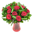 A lush, rounded bouquet of a dozen or more deep crimson roses fills the frame, each bloom open to reveal tightly spiraled, velvety petals that deepen from bright scarlet at the edges to a wine-dark centre. The roses sit in a generous halo of glossy, dark green foliage: smaller, dense leaves-reminiscent of Salal or Italian Ruscus-form a verdant backdrop while broader emerald leaves, shaped and rolled like Aspidistra, are folded into elegant loops and spirals that cradle each head. Those sculpted green arcs lend a contemporary, almost architectural rhythm against the softness of the petals, catching the light in smooth, reflective bands. The stems are gathered in a tall, slender translucent ruby-red glass vase with a subtle ribbed twist that throws delicate vertical highlights and echoes the passionate hues of the flowers. The vase's curved waist and slightly flared lip create a confident silhouette that balances the arrangement's volume. Soft, even studio lighting from the front brings out the roses' velvety texture and casts gentle shadows beneath the leaves, while a crisp white background isolates the bouquet so colour and form read clearly to sight and touch. Close your eyes and imagine a faint, classic rose scent rising from the closely clustered blooms-the kind of fragrance that suggests anniversary evenings and slow, meaningful conversations. This arrangement feels both timeless and edited, perfect as a romantic gift, a refined centerpiece for a special dinner, or a styled photograph for a florist's seasonal showcase in Epping and the surrounding towns.