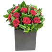 In this elegantly modern composition, deep crimson roses take centre stage, each bloom opening in velvety spirals that read as both intimate and dramatic. The roses are compactly clustered, their layered petals catching the light so that the crimson appears almost satin-sheen against a backdrop of glossy green foliage. The leaves have been artfully curled and folded, forming overlapping ribbons of vibrant emerald that add sculptural movement and a tropical freshness to the design. The flowers sit firmly in a matte black, square box vase whose clean geometry and low profile create a contemporary counterpoint to the softness above; the vase's textured finish absorbs light, making the red blooms glow all the more vividly. Lighting in the shot is clear and modern-cool, steady highlights run across the leaves and along the outer petals, suggesting a bright studio or morning shop window on Epping High Street. If you lean in, you imagine the light, fresh scent of cut leaves mingling with the classic, heady perfume of rose, clean and uplifting rather than heavy. As a local floral design, this arrangement reads as a confident expression of passion and thoughtful restraint-perfect for a modern flat near the station, a romantic anniversary, or a thoughtful gift to mark a special moment. The craftsmanship is visible in the neat, deliberate folds of the foliage and the way each rose is placed to create rhythm and depth; it's the kind of piece that transforms a simple table into an occasion and leaves a lasting, tender impression on anyone who catches its scent.