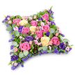 A sumptuous floral pillow, designed as a sympathy tribute, is shown resting against a clean, white background, its soft, cushion-like rectangle gently rounded at the edges to evoke quiet rest. The surface is completely covered with roses in a comforting palette of pinks, creams, and soft yellows: deep fuchsia blooms bring vibrancy, while pastel and buttery tones soften the overall effect. Many of the roses are open wide, revealing intricate whorls of petals that invite the eye to linger. Interspersed among them, deep purple lisianthus flowers curve gracefully, their layered, slightly ruffled petals adding a velvety richness and introducing a calm, contemplative note. Smaller spray roses in pale pink and clusters of tiny lavender filler flowers, much like statice or limonium, are tucked into every available space, giving the design a plush, full appearance with delicate detail at close view. Surrounding and weaving through the arrangement, soft, round eucalyptus leaves in a silvery green hue sit alongside darker, slender, fern-like foliage, the mixed greenery creating contrast, structure, and a sense of natural movement. The lighting is bright yet gentle, casting a faint shadow beneath the tribute, so the colours appear fresh and true, as if the cushion has just been completed for a same-day funeral delivery in Epping. The finished piece feels both elegant and deeply personal, the kind of floral cushion families might choose to place beside a coffin, at a crematorium service, or at a graveside to honour a loved one with grace, tenderness, and enduring remembrance.