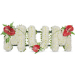 A dignified three-dimensional tribute reads MOM in large, freestanding letters set against a plain white backdrop. Each letter is lovingly composed of dense clusters of small, pristine white blooms-likely spray chrysanthemums or compact carnations-tightly packed to form a soft, cushion-like surface that looks almost cloudlike to the touch. The overall shape is held by a subtle dark green frame that shows at the edges, giving the letters clean lines and quiet structure. To introduce warmth and a heartbeat of colour, three glossy red anthuriums punctuate the design: one placed on the upper left corner of the first 'M', another nestled low in the central 'O', and a third mirrored on the upper right of the second 'M'. The anthuriums' waxy, heart-shaped spathes and central yellow-white spadices offer a tactile contrast to the powdery chrysanthemums, their deep red suggesting devotion and enduring love. Delicate sprays of fern and broader dark green leaves surround each anthurium, the feathered fronds softening the geometry and adding a woodland freshness, while long, slender blades of grass or arcing stems weave between the letters to visually unite the composition. Gentle daylight in the photograph brings out subtle shadows and the sheen on the anthuriums, and there is an implied, faint green-stemmed scent at the edge of the arrangement-clean, vegetal, and respectful rather than overpowering. This is a classic sympathy tribute, designed to convey comfort, honour, and tender remembrance for a mother at a funeral, wake, or graveside service; the restrained white and vivid red together speak of both peace and the warmth of lasting affection.