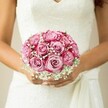 Embodying the spirit of a joyful Epping bride, this bouquet rests gently between warm, lightly sun-kissed hands, each nail finished in a subtle, blush-toned sheen. The arrangement itself is an artistic marvel, composed with devotion reminiscent of mornings in the Rose Garden at Epping's Copped Hall. Plump roses, nearly a dozen, blend gradients of pale pink and vivid fuchsia along each spiralled petal-the colours shifting and dancing as light from a nearby window sweeps across the gown's ornate lace bodice. Wispy sprays of baby's breath are interwoven throughout, diffusing light and evoking a soft, cloudlike innocence that envelops the scene in serenity and romance. A flowing white sash grazes the bride's fingers, adding a hint of movement and grace. The gently blurred backdrop allows the bouquet's artistry-carefully bound hand-in-hand with Epping's floristry traditions-to speak volumes about celebration, hope, and love's quiet anticipation. It's an arrangement that tells a story, making it the perfect reflection of the heartfelt connections and lifelong promises sealed in the heart of Essex.