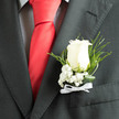 In soft daylight that might filter through a window of our workshop near Epping Station, an ivory rose boutonnière sits proudly against a smart charcoal jacket, the contrast sharpened by the checked texture of a shirt and the confident flash of a red silk tie. The rose's velvety petals unfold with a quiet strength, their creamy tones catching the light in those delicate, living gradations that suggest loyalty and calm. Sprays of baby's breath gather at its base like tiny stars, their purity tempering the boldness of the tie and lending an air of innocence to the composition. Fine slivers of fern and twisted blades of foliage arc around the flower, introducing graceful movement and a darker green that frames the bloom with refined contrast. A slim white ribbon, tied with practiced care, grounds the stems and gives the piece its ceremonial silhouette. Close-held, the boutonnière exudes a cool, fresh scent - the soft rose perfume mingled with the green, slightly earthy note of recently cut leaves - a fragrance familiar to morning markets along the High Street in Epping. Crafted by Flowers Epping with attention to proportion and wearability, this accent is designed for the groom or a loved one who prefers understated elegance: it reads as timeless and romantic, an intimate statement that balances tradition with contemporary tailoring and the quiet pride of a local celebration.