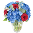 A radiant, hand-arranged bouquet sits in a clear glass vase on a pristine white surface, the composition smelling faintly of cut stems and cool eucalyptus. Dominating the arrangement are large, sky-blue hydrangea clusters - each head made of many delicate, overlapping florets that form a plush, cloud-like mass. Their saturated blue reads like a summer sky after rain, soft to the imagined touch and full of volume. Threaded through these billowy hydrangeas are three deep red roses, their velvety petals tightly spiralled and deliberately placed so each one draws the eye like a punctuation of passion. Tiny white sprays of baby's breath (Gypsophila) float between the bigger blooms, adding an airy, starry texture that lightens the overall weight and creates gentle visual breathing room. Muted, rounded eucalyptus leaves and other fresh green foliage are woven through the bouquet, their cool sage tones and smooth surfaces creating depth and a subtle herbal counterpoint to the sweetness of rose. The clear vase, with a slightly rounded base and tapering neck, shows green stems submerged in water, emphasising the bouquet's freshness and structural balance. Bright, even lighting highlights the saturated blues and crimson contrasts while casting soft, natural shadows that define petal edges. This arrangement feels celebratory and quietly romantic at once - perfect as a table centrepiece, an anniversary gift, or a patriotic display for local gatherings. I often imagine placing a version of this on a window shelf looking out over Epping Forest, where the blue blooms would echo the open sky and the roses would read as an intimate, heartfelt note.