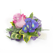 A delicate hand-sized corsage photographed on a clean white background, its composition the kind of piece I often pin for proms and intimate weddings in Epping. At the heart sits a single rose, petals unfurling from a soft lavender-pink center into a more saturated fuchsia at the outer edges, the velvety curves catching a soft, diffused light. Nestled to the right is a tight cluster of indigo-purple florets - hydrangea-like in form - each tiny bloom bearing a bright blue and lime-yellow-green eye that flashes like a garden jewel. Surrounding these blooms is an artful collection of greenery: broad variegated ivy leaves with deep emerald centres and lemon-gold margins that frame the flowers, slender silvery-green foliage suggestive of lavender or dusty miller, and wispy asparagus-fern tendrils that add weightless movement. A single striped green-and-cream blade is curled into a sculptural accent, offering a modern, graphic line. Beneath the arrangement, a dark purple web-like mesh peeks through, creating depth and a contemporary shimmer against the white backdrop. To the lower right, a short strand of luminous white pearls is visible, hinting at a wristlet or classic lapel detail and lending a touch of tradition. The overall mood is romantic and carefully composed - tactile, scented with the faint perfume of rose, and suited to milestones from a ceremony beneath Epping Forest trees to a graduation on the High Street. The piece reads as quietly celebratory: crafted to be pinned, admired up close, and remembered.