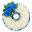 A bespoke funeral tribute shaped as a perfect '0', photographed from directly above against a clean white backdrop. The foundation is an unbroken ring of tightly clustered white spray chrysanthemums-hundreds of tiny florets pressed close together to form a cloud-soft, velvety surface that reads as serene and pure. The chrysanthemum petals fan outward in subtle layers, catching a gentle, diffuse light that gives the wreath a soft, matte glow and suggests a faint, green-tinged sweetness in scent. Where the upper-left curve meets the outer edge, three royal blue roses gather as a deliberate, vivid focal point; their petals are rich and velveteen, unfurling from neat, tight centres into full, sculpted blooms. Deep glossy foliage nestles behind the roses, the dark green leaves offering a cool, living contrast that frames the blue trio and anchors them against the white. A pleated royal blue satin ribbon trims both the inside and outside of the number, its ruffled edge creating a scalloped silhouette that echoes the roses' intense hue and lends the tribute an elegant, formal finish. The overall feeling is dignified and contemplative-balanced geometry softened by texture, colour and careful shading-suitable for a memorial service, a milestone age tribute, or a ceremonial farewell. As a local touch, this style is one we often prepare for families in Epping and nearby villages when they request a personalised number wreath for a service at St John's or a remembrance held near the town centre; the cool blue and pure white read as a peaceful salute, quiet and resolute.