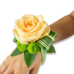 A delicate wrist corsage rests against the inner forearm, set on a clean white background so every texture reads clearly; the skin tone here is a warm, light to medium brown that gently cradles the arrangement. The focal flower is a single, large rose in a soft peach-apricot shade, its many velvety petals uncoiling toward the centre in a perfect, romantic spiral that seems to hold a quiet breath of fragrance. Beneath and to one side, two vivid lime-green button chrysanthemums-tight, spherical pom-pom blooms-provide a playful, tactile contrast: their tiny, clustered florets look almost like confetti against the rose's smooth surface. A rich foundation of glossy dark-green leaves, likely ruscus or salal, fans out and gives the corsage depth and structure, while a distinct variegated leaf, striped with pale streaks, is expertly folded and curled to create a clean, architectural sweep that points away from the wrist and adds modern movement to the design. The arrangement sits as if ready for a hand to lift in greeting; an elastic or ribbon holds it unseen beneath the foliage. Soft, natural light highlights the rose's warm tones and the chrysanthemums' electric green, and there is an implied scent of rose warmed by morning air. Perfect for a wedding morning, prom or intimate celebration in Epping - whether beneath the trees of Epping Forest or on the High Street - this corsage feels both handcrafted and timeless, a small wearable bouquet that promises remembered moments and quiet elegance.