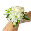 A single wrist corsage captured against a clean white backdrop, resting on a light-skinned wrist and composed with the calm restraint of a seasoned florist. At its heart sits a creamy ivory rose, petals unfurling in velvety layers that catch soft light and read as pure, muted warmth. Just below and slightly to the left, a larger trumpet-shaped white bloom-reminiscent of Alstroemeria or a delicate lily-opens wide, its throat tinged a subtle green-yellow that gives the composition a gentle centre glow. Scattered through the design are tight green buds that promise further bloom, and tiny airy sprigs of white Gypsophila which lend the corsage a cloud-like softness and visual breath. Near the wrist a small daisy-like head with a bright yellow centre adds a cheerful punctuation, its texture crisp against the rose's velour. Broad, glossy deep-green leaves frame the flowers, their waxy surfaces providing a cool contrast to the warm ivory tones and helping the arrangement sit naturally on the skin. The whole cluster is secured on an elegant multi-strand pearl bracelet: uniform, luminous pearls thread together to form a soft, elastic band that encircles the wrist and elevates the piece into classic formality. Imagine the faint, clean scent of fresh blooms and a barely-there powdery rose note when the wearer moves. This wrist corsage reads as timeless and refined-perfect for proms, wedding parties, or any quiet rite of passage-and shows careful craftsmanship and balance in both floral choice and placement, the kind of piece one would trust a local maker to create with steady hands and a thoughtful eye.