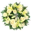 A quietly dignified circular wreath, arranged flat and photographed from above against a clean white background, where creamy ivory roses and soft pale yellow lilies nestle into a deep, verdant cradle of foliage. The wreath is densely packed so each bloom sits close to the next: roughly a dozen to twenty fully opened roses reveal their tightly furled, satin-like petals in warm cream and ivory tones, while the lilies-large, trumpet-shaped flowers in a gentle lemon-petal yellow-are interspersed in varying stages from closed bud to fully unfurled blossom. Feathery fern fronds fan out beneath and between the flowers, giving the piece a delicate, lacy edge, and smaller rounded sprigs of eucalyptus-like leaves add cooler, grey-green notes and a subtle matte texture. Soft, even daylight in the photograph catches the roses' inner spirals and the lilies' graceful curves, and if imagined in person the wreath would offer a faint, comforting scent-powdered sweetness from the roses and a green, slightly citrus lift from the lilies. The circular form reads as a quiet symbol of remembrance and continuity, appropriate for a funeral or memorial setting, whether placed on a chapel table, carried to a graveside, or laid at the foot of a photograph during a service. As a local florist working across Epping-from the High Street to the edges of Epping Forest-we craft this selection to convey a serene message of peace and heartfelt condolence, its palette of cream, soft yellow and varied greens offering a gentle reassurance in moments of loss.